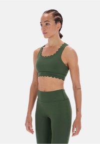 Zielony sportowy biustonosz z falistymi brzegami, wykonany z gładkiej tkaniny. W zestawie z wysokim stanem, dopasowanymi zielonymi legginsami. Gładka tekstura, dopasowany krój.