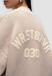 Jasno beżowy sweter z dzianiny z haftowanym napisem "WRSTBHV" i "030" w kremowym kolorze, z teksturowanym materiałem i żebrowanymi detalami.