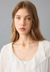 Bílá halenka s volánky a mašlí u krku, vrstvená látka a měkká textura. Modelka má dlouhé, vlnité vlasy a nosí kulaté náušnice.