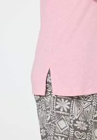 Camiseta rosa claro de manga corta con aberturas laterales, superpuesta sobre pantalones grises con patrones que presentan diseños geométricos y acentos florales.