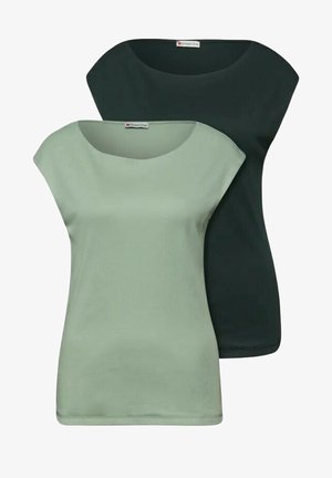 Deux femmes portent des tops unis à manches courtes en vert sauge et vert foncé avec des jeans en denim, l'une accessoirisée d'un foulard à motifs autour du cou.