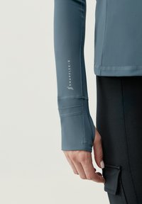 Camiseta deportiva de manga larga en azul pizarra, con orificios para pulgares en los puños, textura suave y marca reflectante en el brazo.