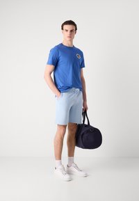 Mann steht in blauem T-Shirt und hellblauen Shorts, hält eine marineblaue Reisetasche, trägt weiße Sneakers und Socken vor einfarbigem Hintergrund.