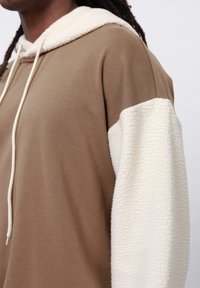 Felpa con cappuccio marrone caratterizzata da una manica in tessuto sherpa crema e un corpo liscio, con lacci bianchi e vestibilità rilassata.