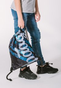 Batoh na stažení v černé barvě s modrým a stříbrným grafickým designem, s velkým logem a texturovanou tkaninou. Uživatel má na sobě černé tenisky.