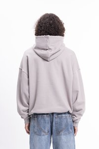 Persona con capelli ricci indossa un ampio hoodie grigio chiaro e pantaloni jeans blu larghi, girata di spalle su uno sfondo bianco.