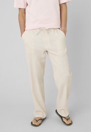 Personne portant un pantalon beige clair à cordon de serrage et des tongs noires, mains dans les poches, debout devant un fond gris uni.