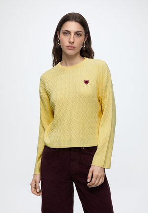 Femme aux longs cheveux bruns portant un pull jaune à torsades avec un petit écusson rouge en forme de cœur et un pantalon marron foncé, debout sur un fond blanc.