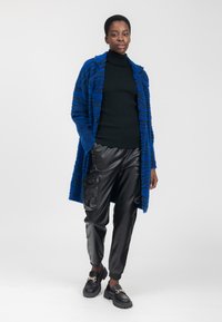 Un cardigan largo de peluche a rayas azul y negro sobre un cuello de tortuga negro, combinado con pantalones cargo de cuero negro y mocasines negros con suelas gruesas.