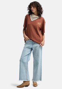 Braunes, übergroßes T-Shirt mit gesticktem Design, weißem texturiertem Kragen, kombiniert mit hellblauen weit geschnittenen Jeans und braunen Cowgirl-Stiefeln.