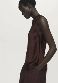 Top bordeaux senza maniche realizzato in un tessuto lucido e strutturato. Presenta un collo alto e una vestibilità rilassata, abbinato a pantaloni scuri coordinati.