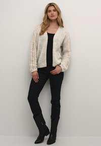 Lightweight, cream-colored knitted cardigan with ribbed edges and open front; detailed, textured sleeves. Worn over a black top and pants, with black boots.