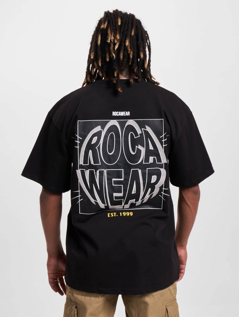 Juoda didelio dydžio marškinėliai su dideliu, ryškiu baltu „ROCA WEAR“ grafiniu piešiniu ant nugaros, su „EST. 1999“ geltonai. Lygus medvilninio audinio paviršius.