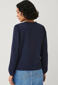 Blusa navy a maniche lunghe con tessuto testurizzato, dettaglio con rouches sul colletto e maniche a sbuffo, abbinata a una gonna di denim blu chiaro.