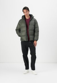 Groene gewatteerde jas met een capuchon, voorzien van een rits en elastische manchetten, gedragen over een bordeauxrood shirt en zwarte broek, gecombineerd met witte sneakers.