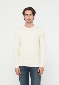 Jasnokremowy sweter dziany z długim rękawem i okrągłym dekoltem, wykonany z miękkiego materiału. Noszony z niebieskimi jeansami.