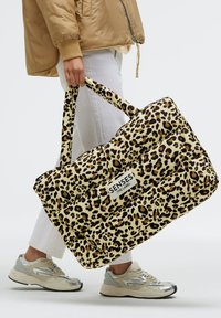 Leopard print tote bag with padded design, cream background, and black/brown spots. Features sturdy handles and a label patch.