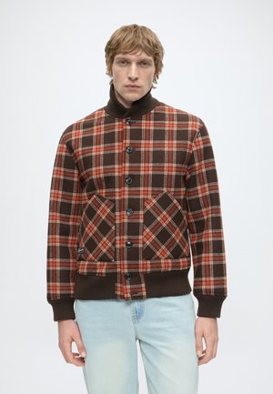 Homme portant une veste à carreaux marron et orange à boutons, avec col, poignets et ourlet côtelés, associée à un jean bleu clair sur fond blanc.