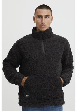Black fleece pullover with a half-zip neck, textured fabric, and a kangaroo pocket. Features a small branded patch at the hem.
