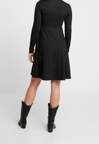 Musta ribbed mekko, jossa on pitkät hihat, liehuva helma ja tiukka vartalo. Päällä polvipituiset mustat saappaat.