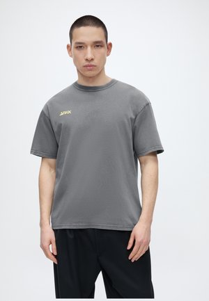 Mladý muž s krátkymi vlasmi, ktorý nosí sivé tričko značky ARKK a čierne nohavice, stojí na bielom pozadí.