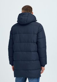Marineblå polstret parka med hette, med quiltede seksjoner og elastiske mansjetter, designet for varme og dekning.