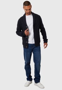 Cárdigan azul marino oscuro con un patrón de punto trenzado, cuello de chal y seis botones, combinado con una camiseta blanca y jeans azules.