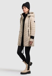 Beige Daunenjacke mit Kapuze, vertikalen Steppungen, schwarzen Akzenten, seitlichen Reißverschlüssen, getragen über einem schwarzen Outfit und beigefarbenen Stiefeletten.