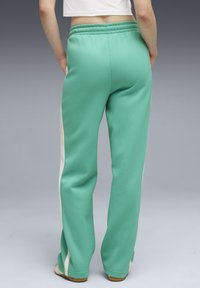 Άτομο που φοράει πράσινη μέντα φόρμα με λευκές ρίγες στα πλάγια, όρθιο με τα χέρια στις τσέπες, μπροστά σε ουδέτερο γκρι φόντο.