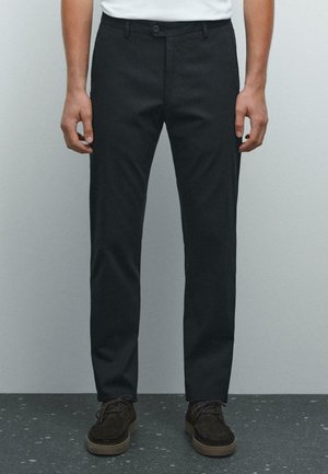 Man wearing slim-fit black trousers and dark brown suede lace-up shoes standing on a speckled floor against a plain wall.