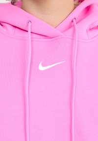 Rožinė viršutinė sukapota iš minkštos medžiagos, turinti priekinius kišenę, raištelius ir baltą Nike logotipą ant krūtinės.