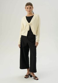Cardigan lavorato a maglia color crema con chiusura frontale a fiocco, abbinato a pantaloni neri a gamba larga a vita alta e tacchi neri con cinturini.