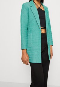 Türkisfarbenes Blazer mit Fischgrätenmuster, einer einzigen vorderen Tasche, Reverskragen und taillierter Passform, kombiniert mit einem schwarzen Oberteil und Hose.