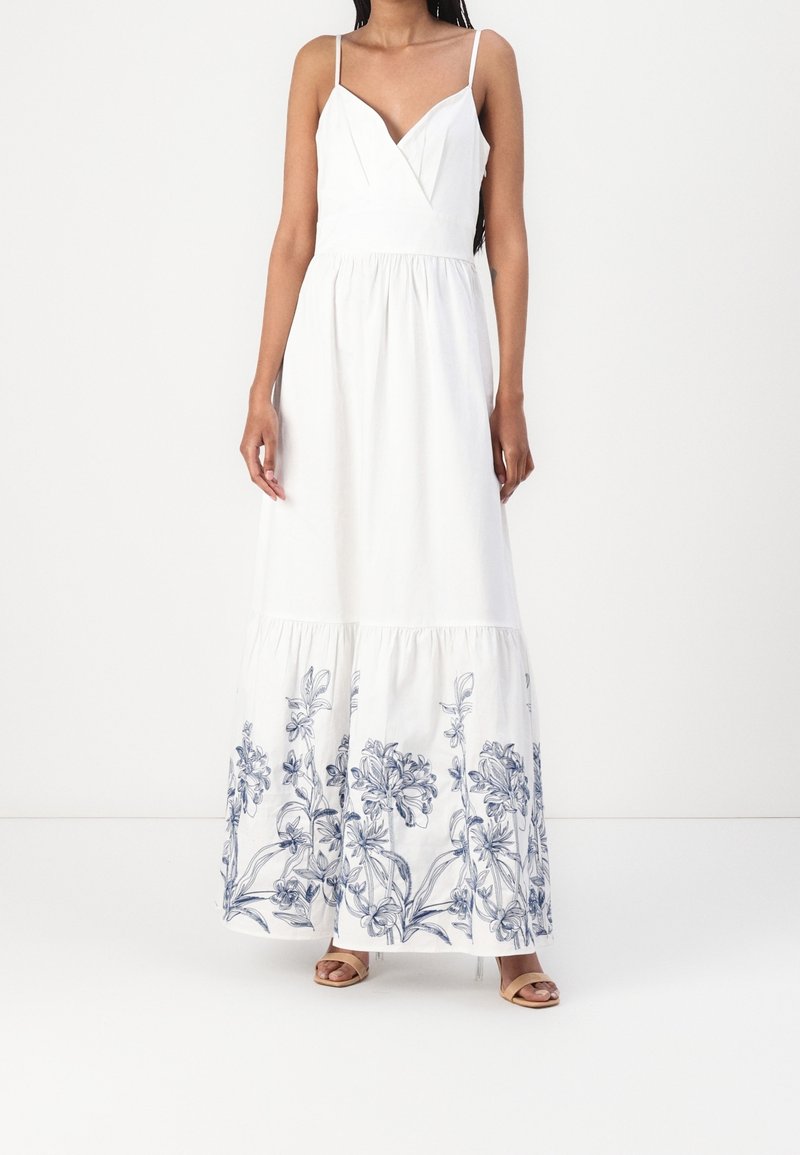 Femme portant une robe maxi blanche sans manches avec une broderie florale bleue près de l'ourlet, associée à des talons ouverts beige.