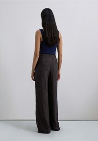 Mujer con cabello largo y oscuro de pie de espaldas, vestida con una blusa sin mangas azul marino y pantalones anchos de tiro alto con un estampado marrón.