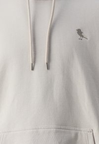 Hellgraue Kapuzenjacke aus weichem Material mit einer Kängurutasche, Kordelzügen und einem kleinen Vogel-Logo in einem dunkleren Grau.