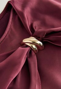Burgundy satin stof med en glat overflade, prydet med en snoet guldfarvet metaldetalje ved knuden. Slankt og elegant design.