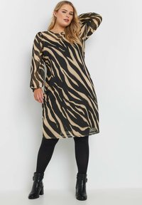 El dress con estampado animal en beige y negro, con mangas largas y un ajuste holgado. Usado con leggings negros y botas de tobillo. Tejido suave y ligero.