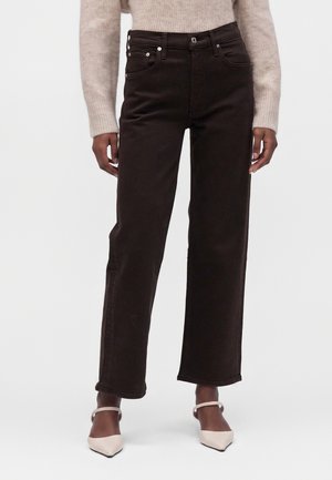 Person iført sorte high-waist straight-leg jeans, beige striktrøje og spidse beige hæle med ankelremme, stående mod en hvid baggrund.