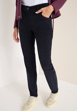 Pantaloni scuri navy con vestibilità slim, tasche frontali e chiusura con bottone. Indossati con sneaker beige chiaro. Tessuto morbido e liscio.