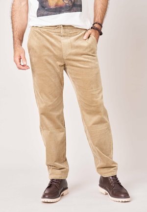 Man die beige corduroy broek en donkerbruine leren laarzen draagt, staat met één hand in zijn zak tegen een effen achtergrond.
