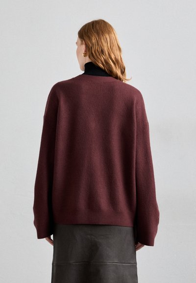 Burgundin värinen ylisuuria neuletta, joka on valmistettu teksturoidsesta neulekankaasta. Tässä on löysä istuvuus ja tippuva olkapäädesign, yhdistetty mustaan hameeseen.
