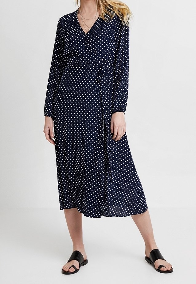 Robe portefeuille bleu marine à pois, manches longues, longueur mi-mollet, et ceinture à la taille. Portée avec des sandales plates noires.