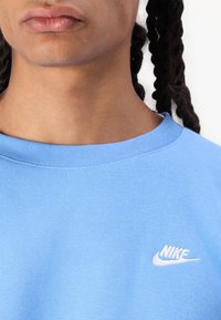 Világoskék Nike pulóver, kerek nyakkal. Bal mellrészen fehér hímzett logóval rendelkezik. Puha textúrájú, normál szabású.