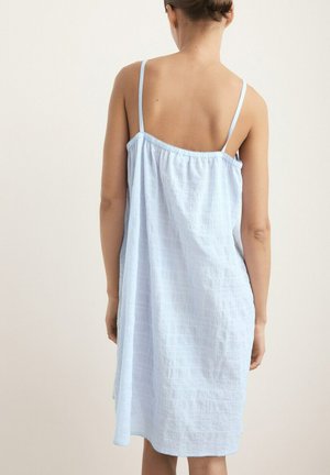 Frau von hinten gesehen, die ein hellblaues ärmelloses lockeres Kleid mit dünnen Schulterriemen vor einem schlichten Hintergrund trägt.