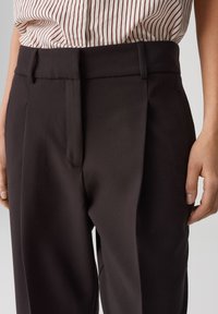 Brown tailored trousers with a smooth texture, featuring front pleats and belt loops, paired with a striped white and red shirt.