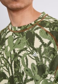 Zelena in kremna srajca z vzorcem kaktusov, teksturirana tkanina in rjavi šivi. Logotip v obliki trikotnika na levi strani.