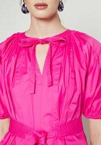 Woman wearing a bright pink dress with puff sleeves, a tied bow neckline, matching belt, and large dark patterned hoop earrings.