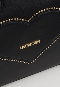 Sac en cuir noir avec surpiqûres en forme de cœur et bordure de perles dorées, présentant une petite plaque logo dorée "LOVE MOSCHINO".