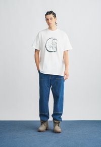 Biele bavlnené tričko s kruhovým grafickým dizajnom, skombinované s modrými rifľami a sivými čižmami s textúrovanou podrážkou na modrom podklade.